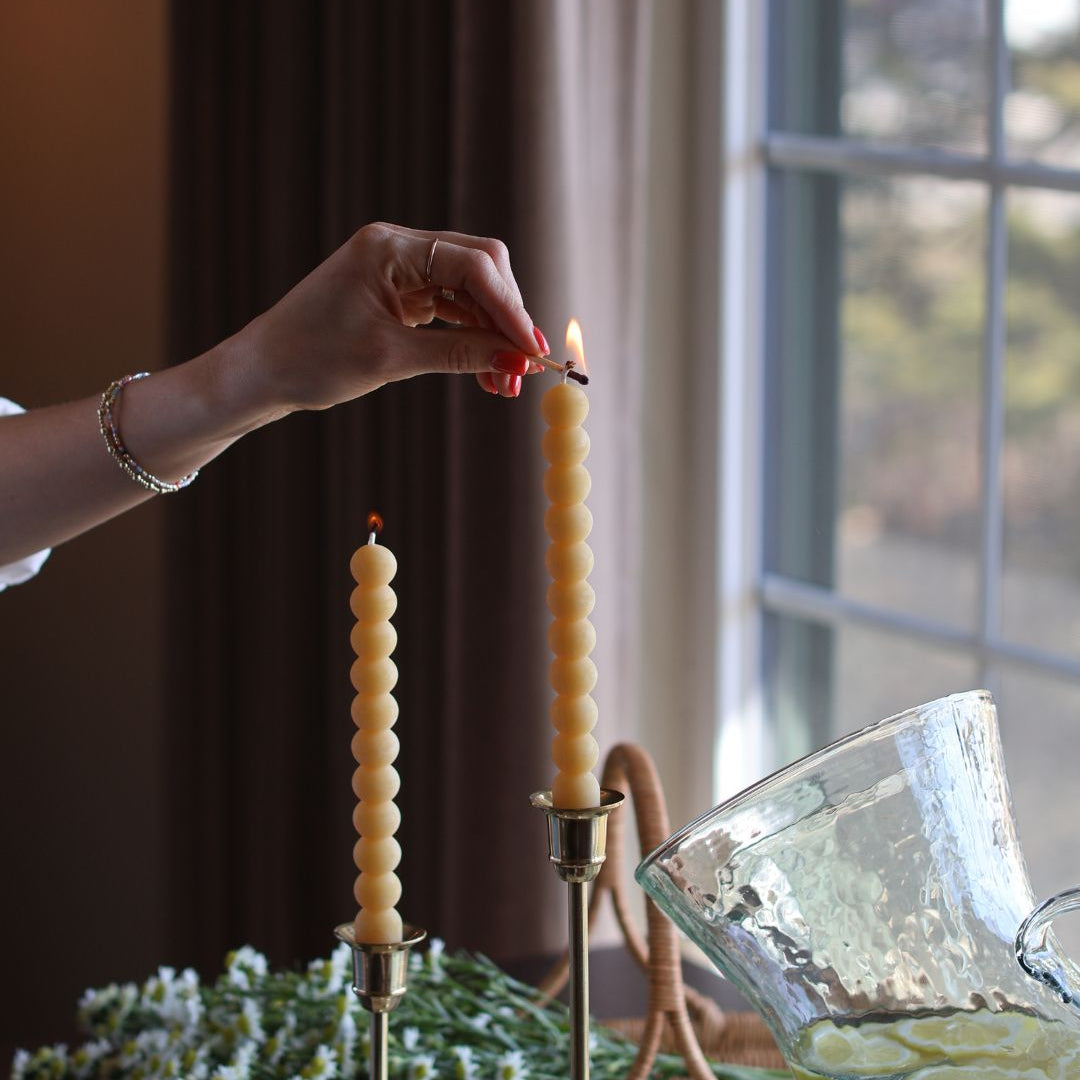 Women's hand using a wooden match to light two locally made beeswax candles resting on golden candelabras. 