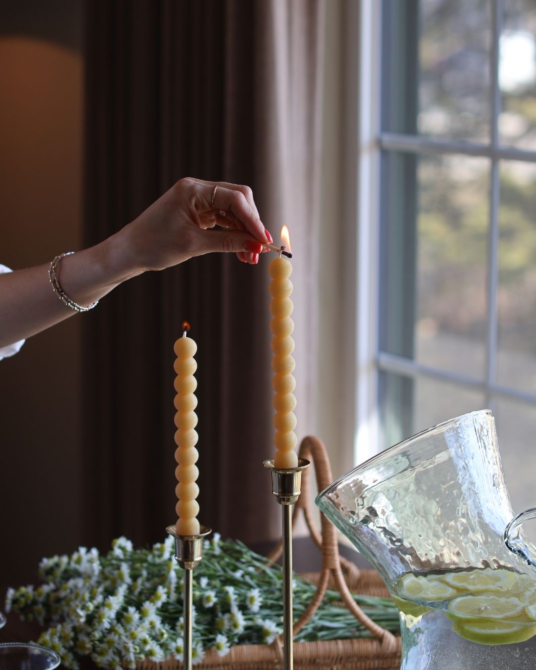 Women's hand using a wooden match to light two locally made beeswax candles resting on golden candelabras. 