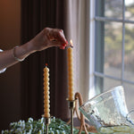 Women's hand using a wooden match to light two locally made beeswax candles resting on golden candelabras. 