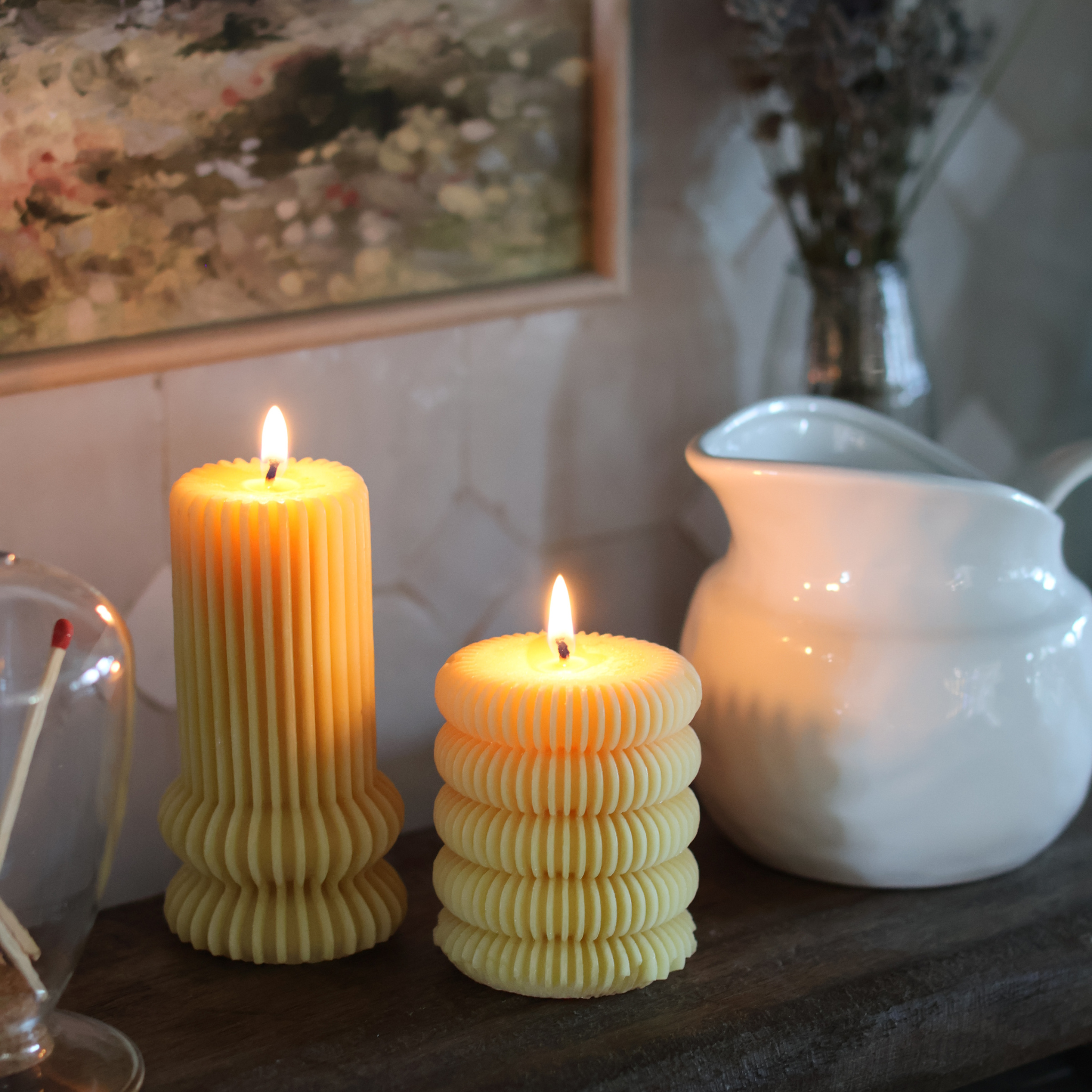 Two ribbed locally made beeswax candles on a wooden surface with a white pitcher and decorative elements in the background.