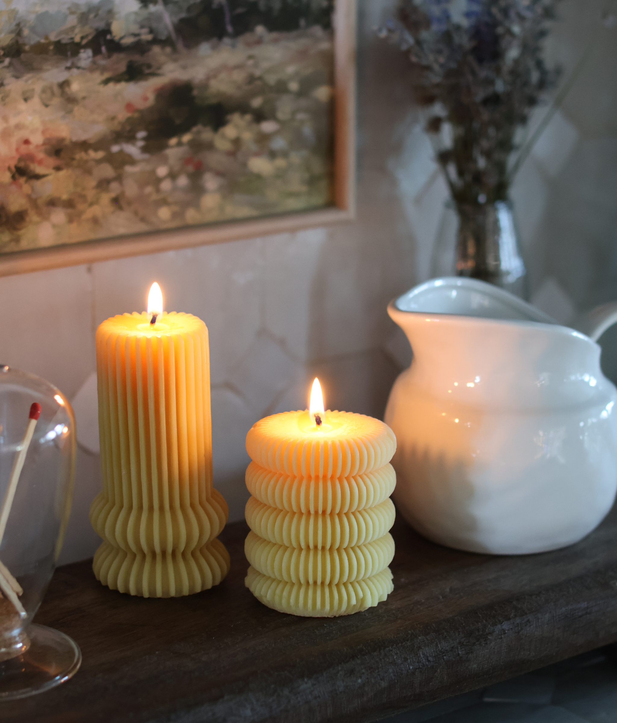 Two ribbed locally made beeswax candles on a wooden surface with a white pitcher and decorative elements in the background.