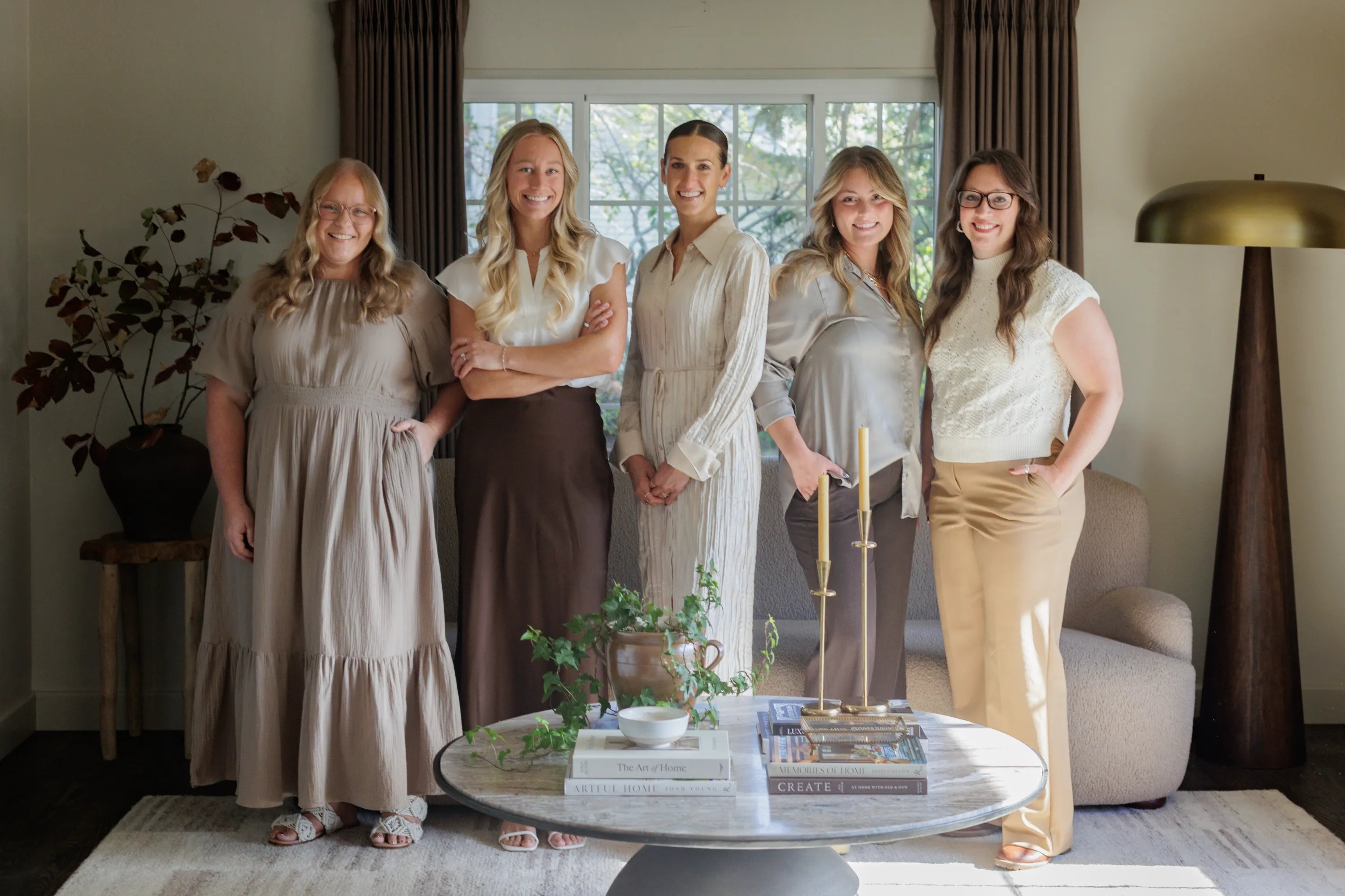 The S. Flynn Home Team, featuring the founder in the middle, accompanied by her store managers and marketing associates in a styled living room with a beige sofa, marble coffee table, decorative books, candelabra, and a gold and wooden lamp. 