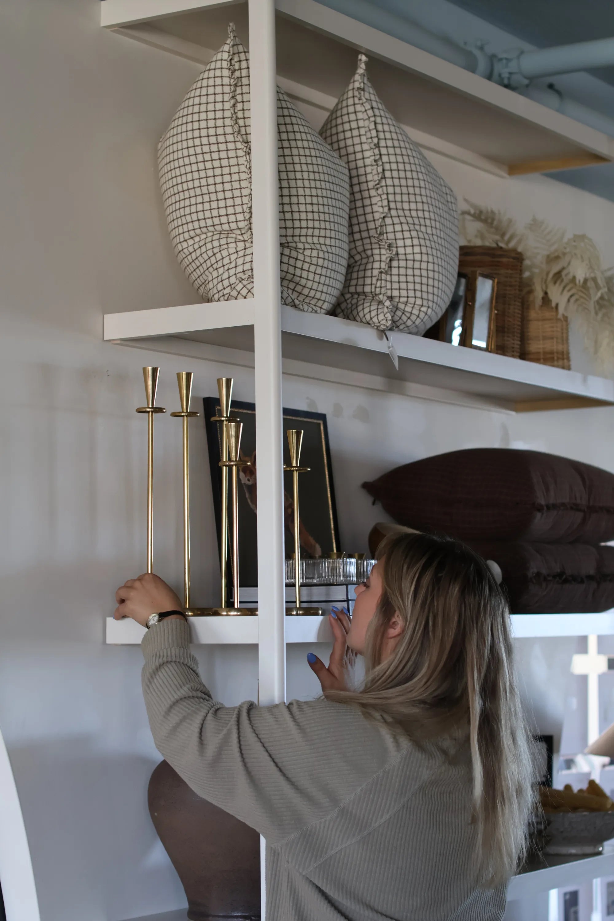 Stockist at S-Flynn-Home store. Adding golden candelabras to a shelf with other decorative items such as throw pillows, artwork, ceramic bowls, and wicker baskets. 