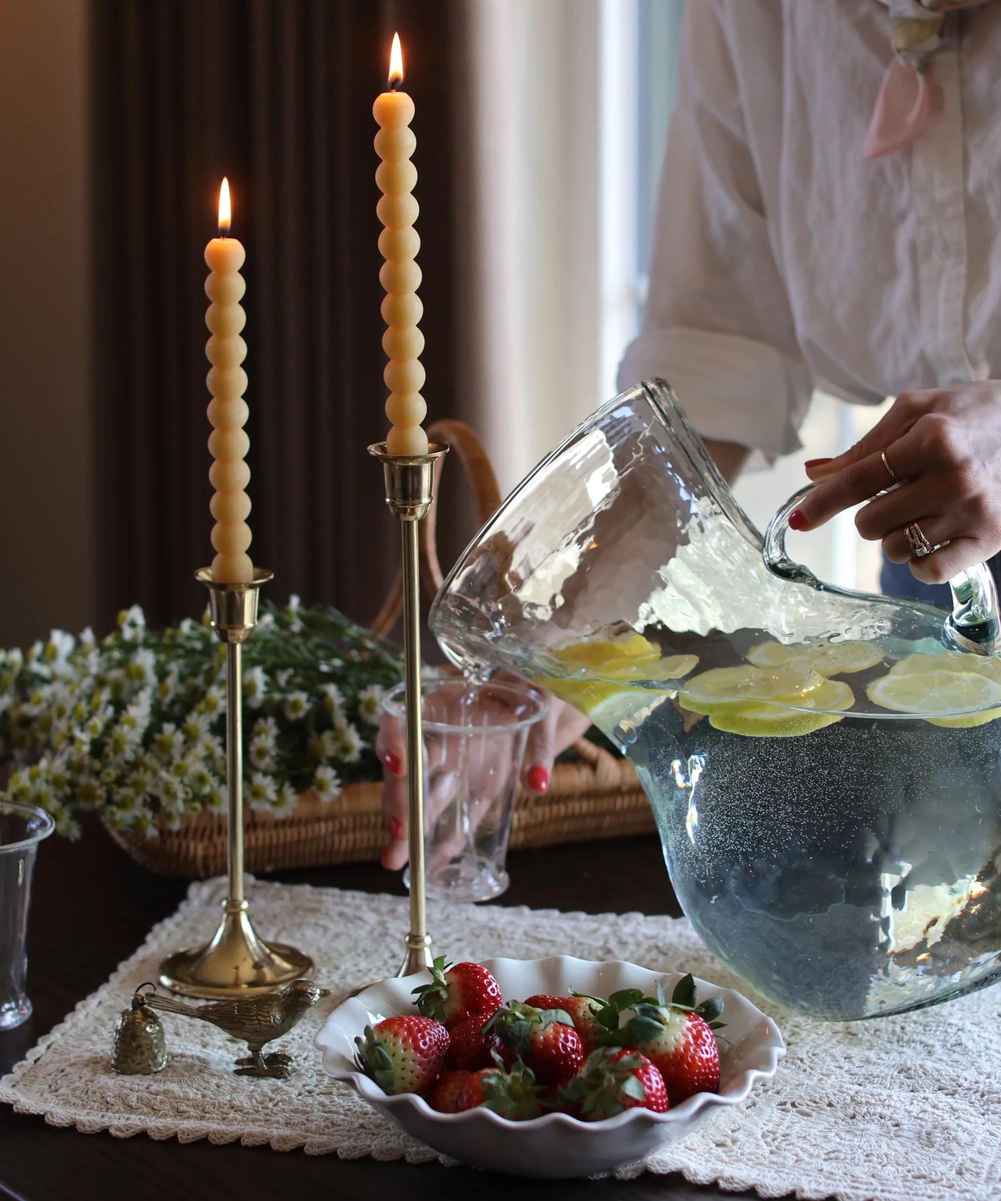 Founder of S. Flynn Home pouring water into a glass cup on a wooden table with two locally made beeswax candles and a scalloped ceramic bowl of strawberries. 